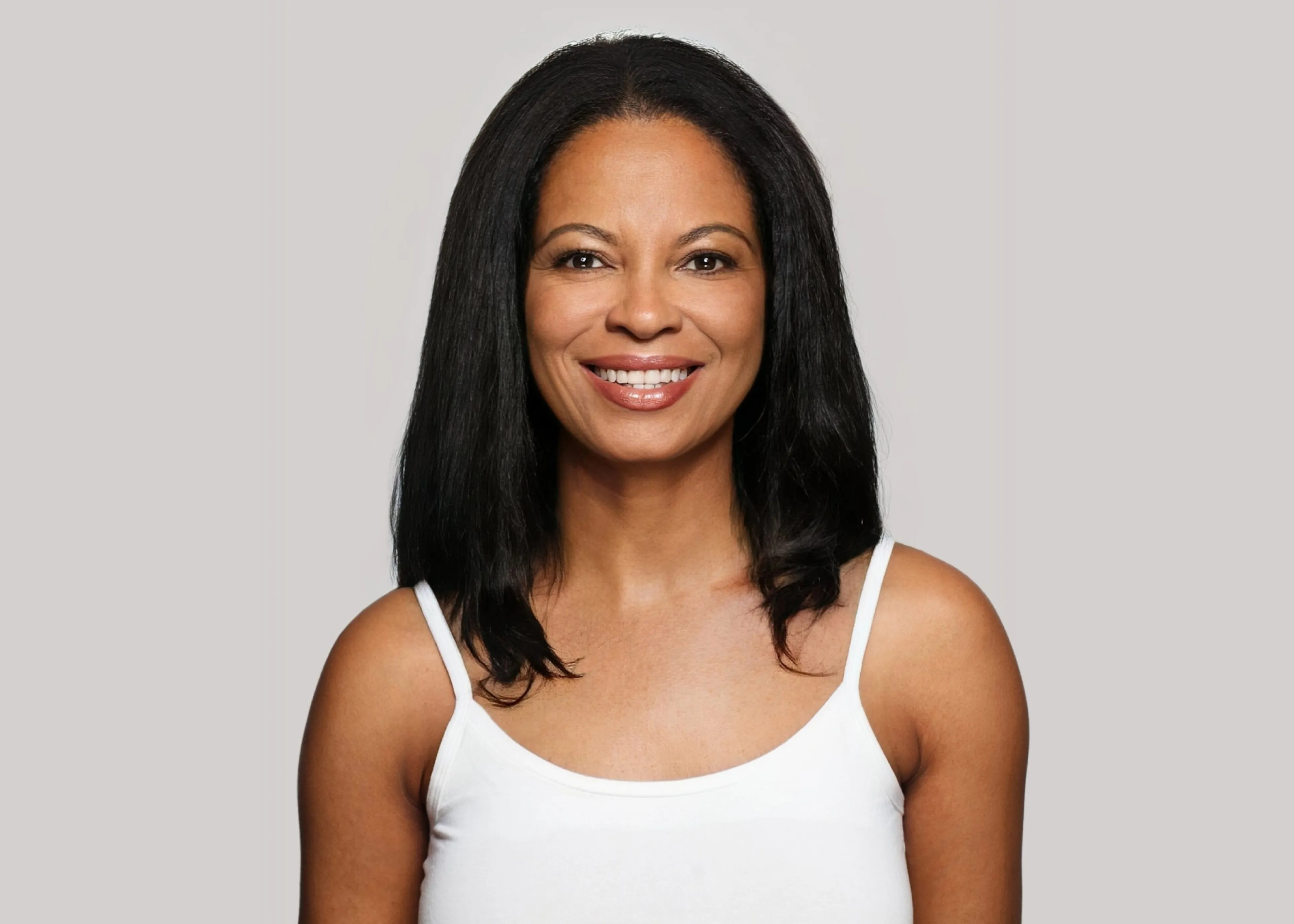 Smiling woman with medium-length black hair wearing a white tank top against a plain gray background.