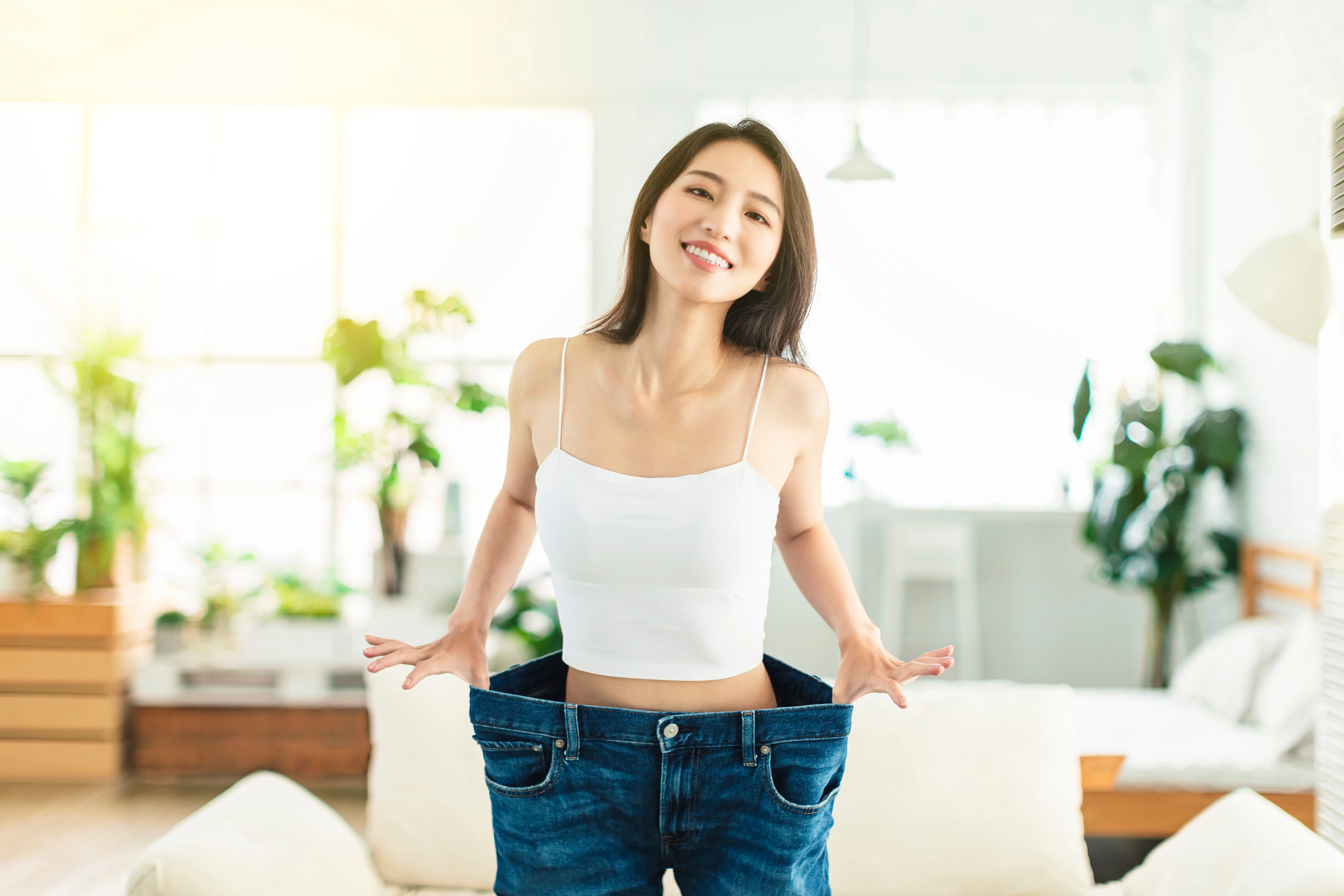 Smiling woman in a white tank top wearing oversized blue jeans, showing weight loss in a bright living room.