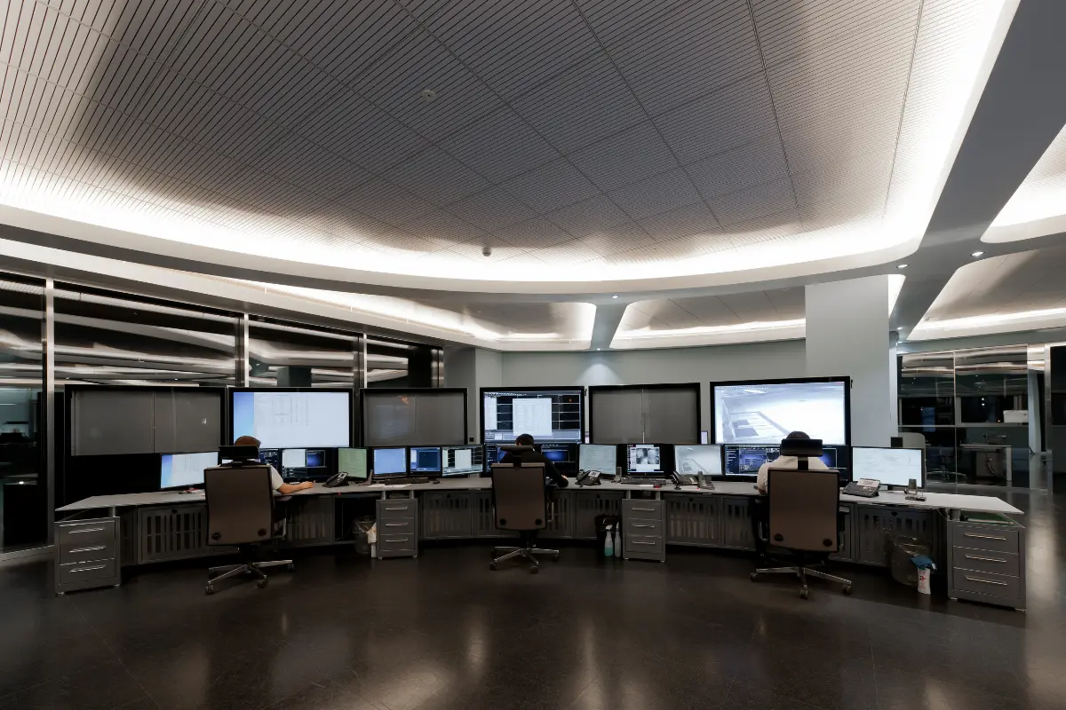 Sala di controllo con tre persone sedute davanti a una fila di schermi di computer in un ambiente moderno e illuminato.