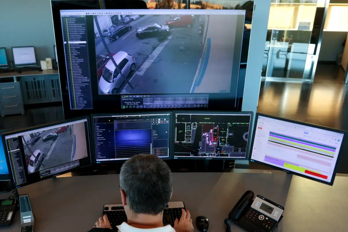 Operatore di sicurezza che monitora più schermi con immagini di telecamere di sorveglianza e mappe digitali in una sala di controllo.