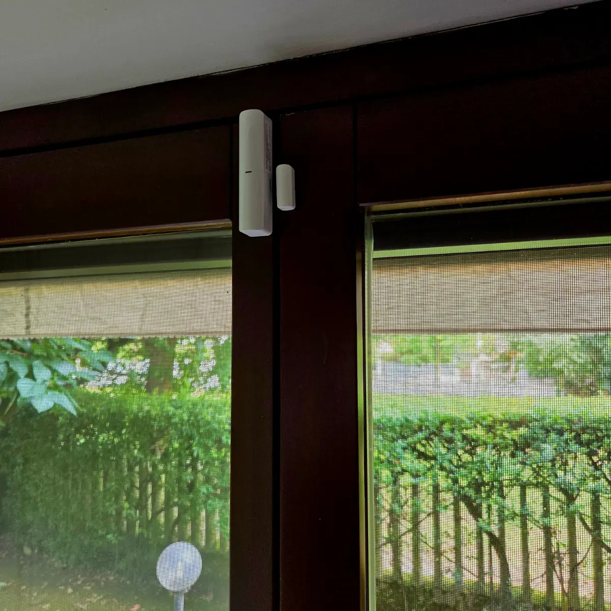 Sensore di apertura porta o finestra bianco montato su telaio di legno scuro con vista su giardino esterno.