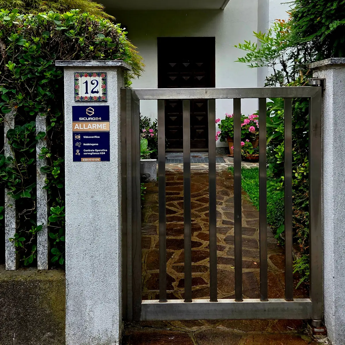 Cancello metallico davanti a un vialetto con pietre irregolari, piante e fiori rosa vicino alla porta di casa bianca.