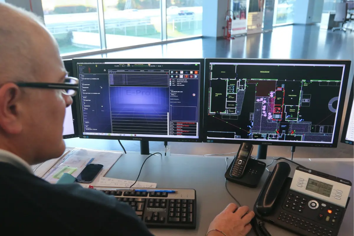 Uomo seduto a una scrivania con due monitor che mostrano software di progettazione e planimetria, tastiera e telefoni da ufficio.