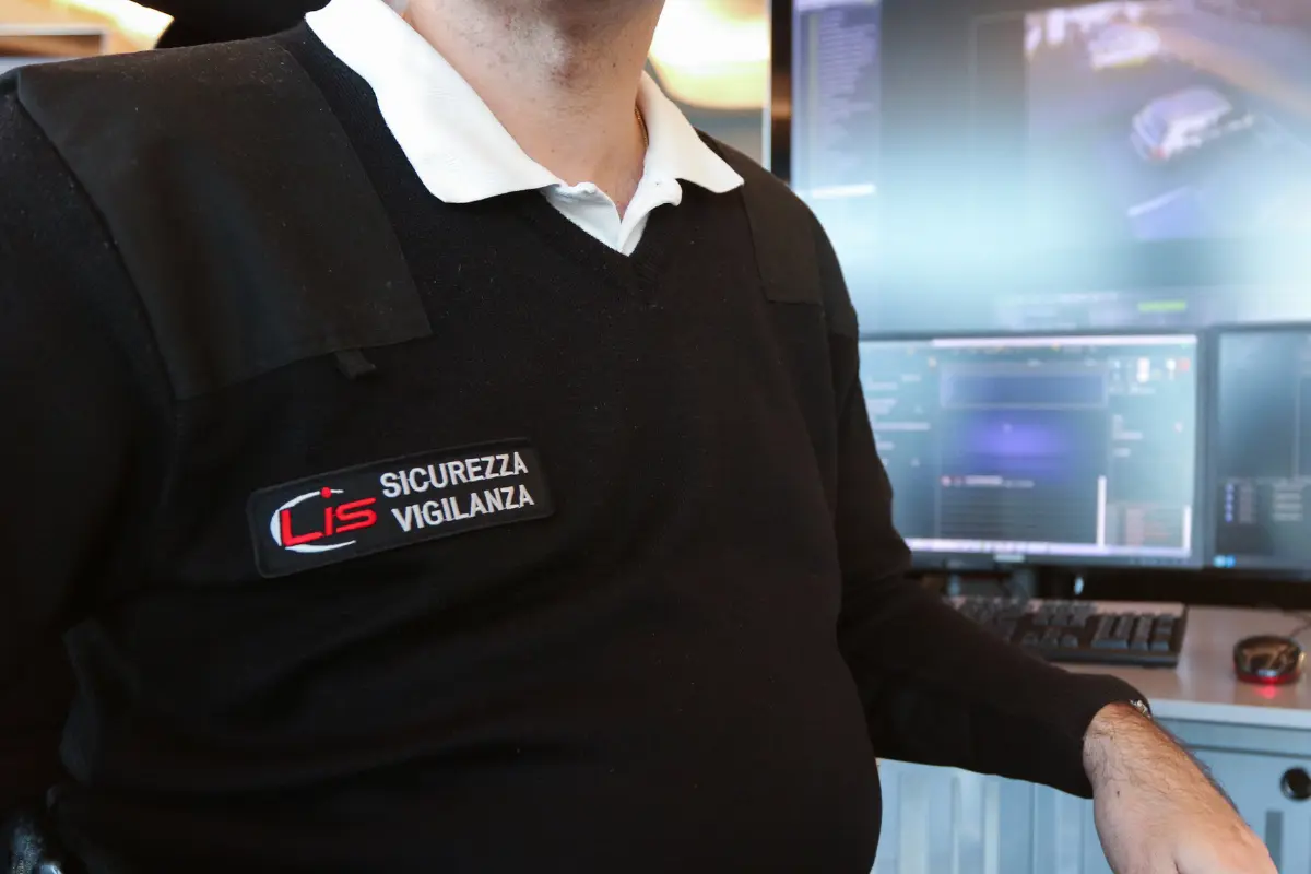 Guardia di sicurezza in divisa nera con targhetta Lis Sicurezza Vigilanza davanti a schermi di controllo.