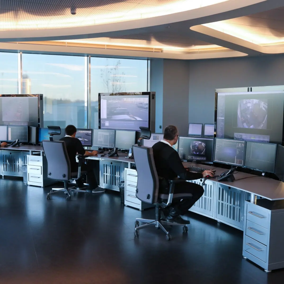 Due operatori seduti a scrivanie con più monitor in una moderna sala di controllo con grandi finestre.