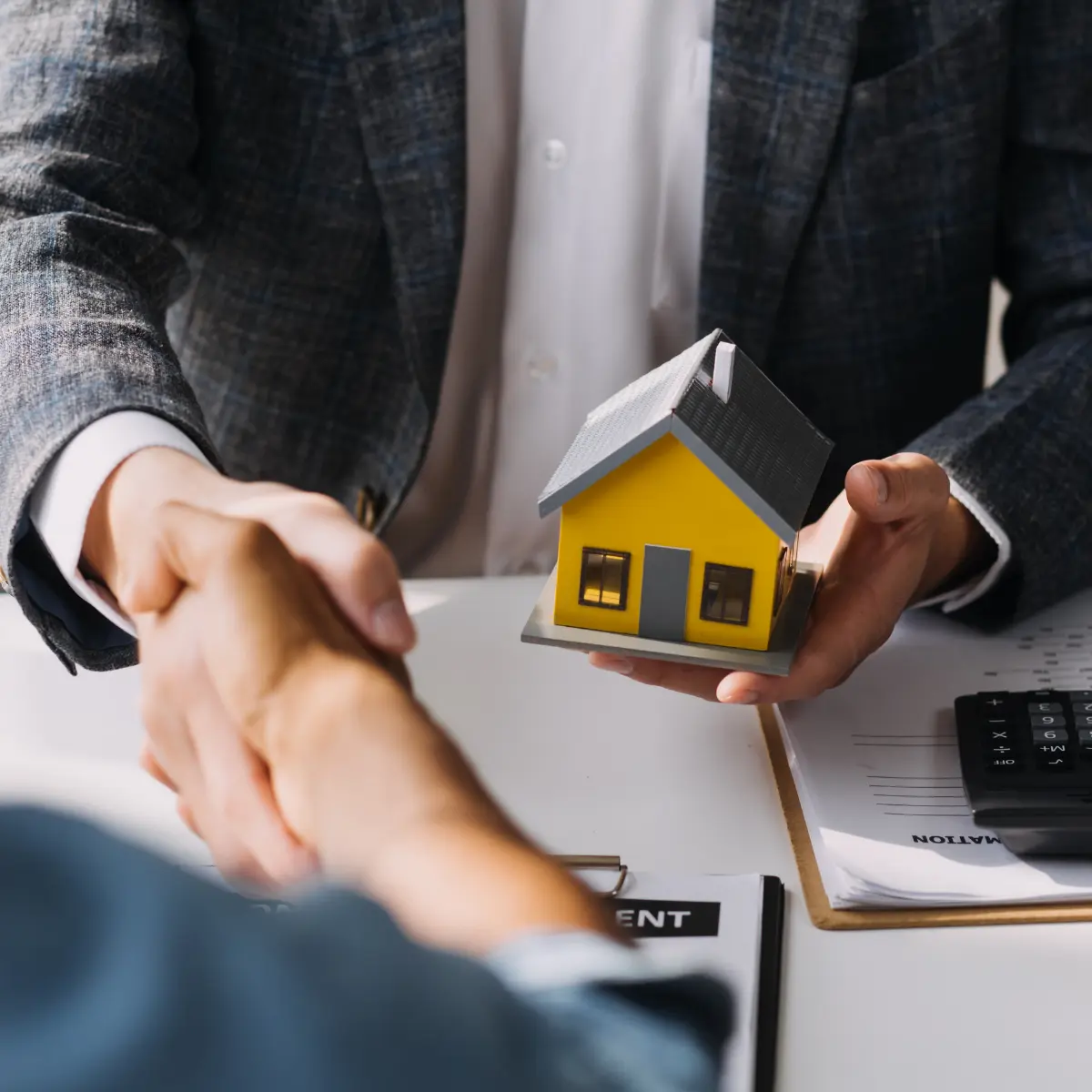 Due persone si stringono la mano sopra un documento con un modellino di casa gialla tenuto da una mano.