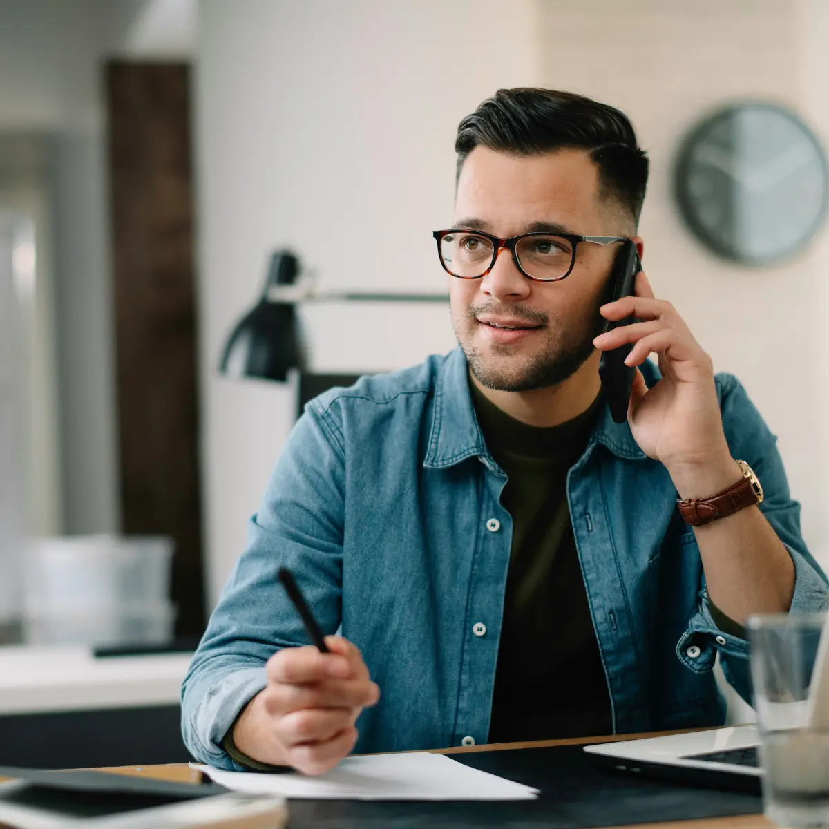 Uomo con occhiali che parla al telefono tenendo una penna sopra dei fogli sulla scrivania.