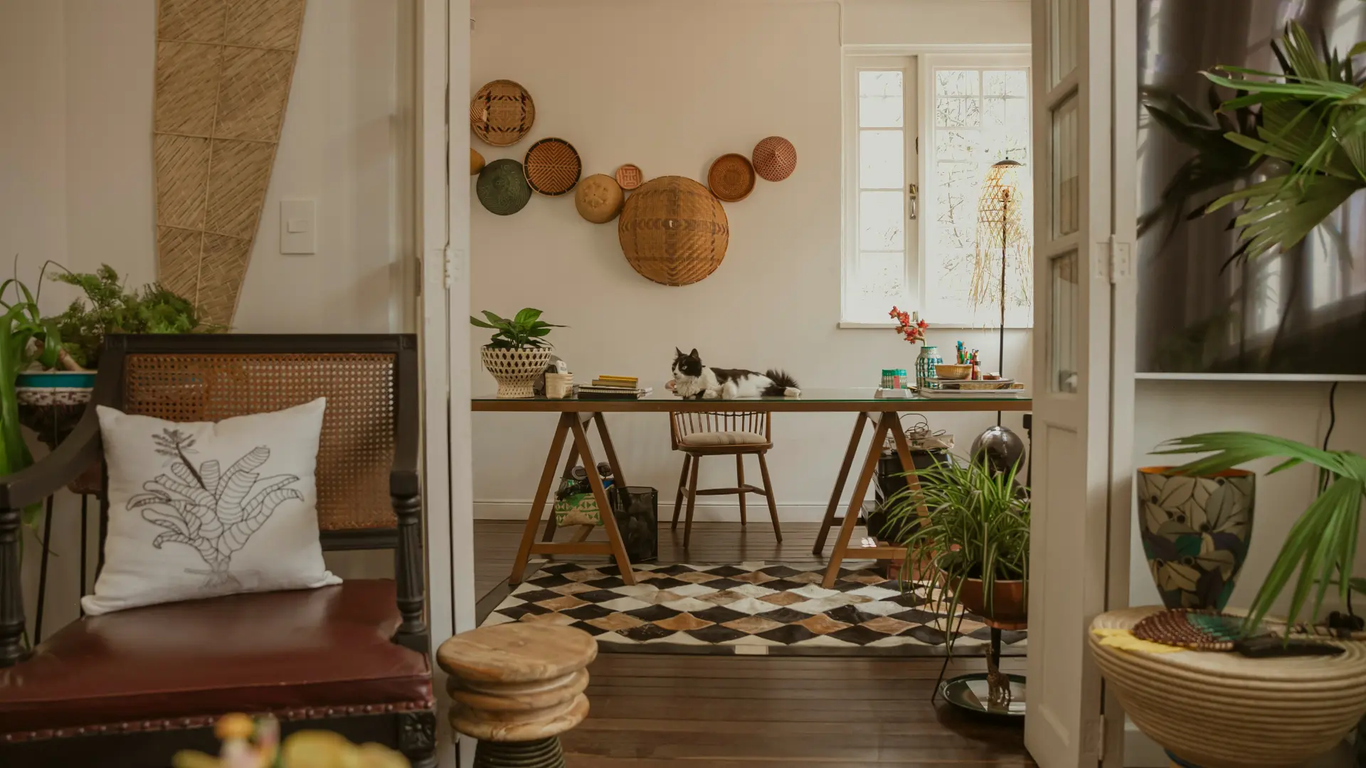 Soggiorno con pavimento in legno scuro, grande tavolo con gamba a cavalletto, tappeto geometrico, gatto nero e bianco sul tavolo, sedie e piante decorative.