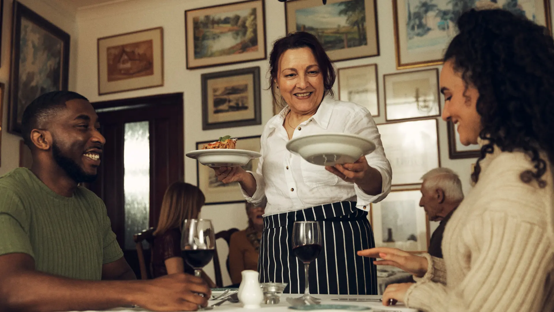 Cameriera che serve piatti a due clienti sorridenti seduti a un tavolo con bicchieri di vino rosso in un ristorante accogliente.