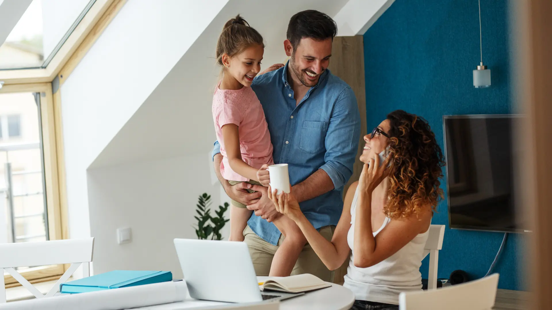 Famiglia sorridente in una stanza luminosa con finestra, una donna parla al telefono mentre un uomo tiene una bambina e le porge una tazza.