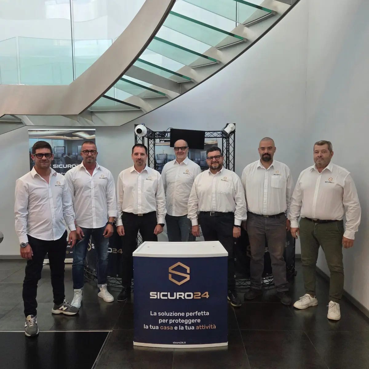 Sette uomini in camicie bianche con il logo Sicuro24 posano in piedi dietro un banchetto con il logo e lo slogan aziendale in un ambiente moderno con scala a vetri.