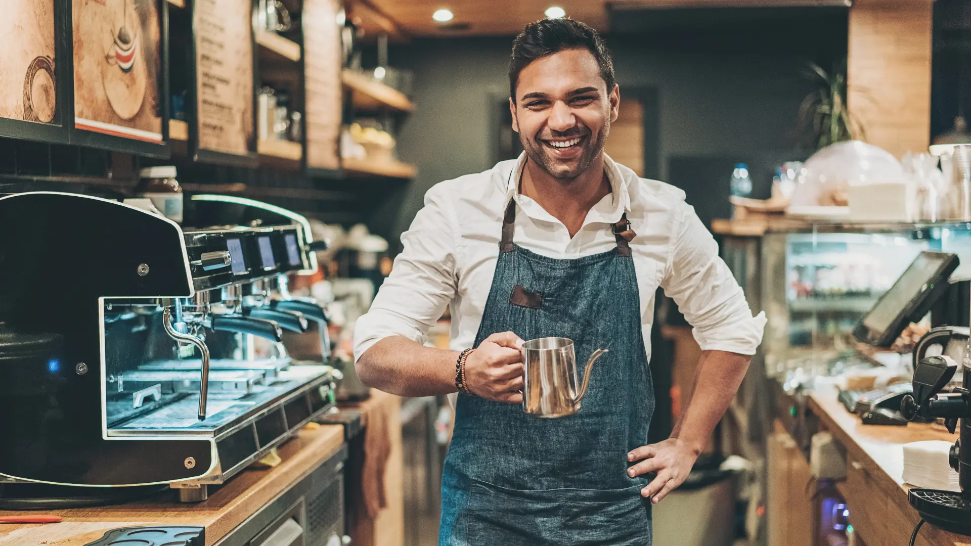 Barista sorridente in grembiule blu tiene una brocca metallica accanto a una macchina per caffè espresso in un bar.