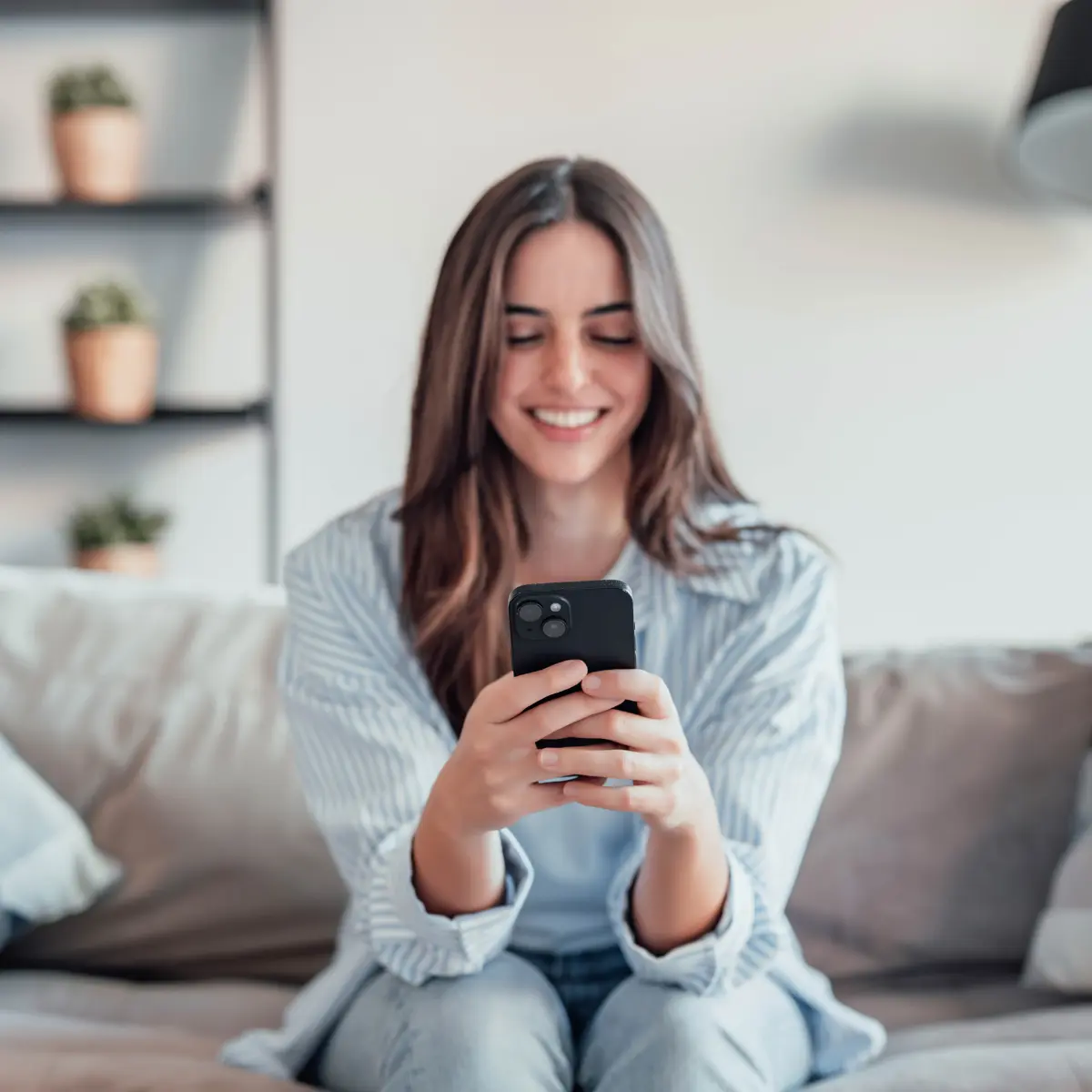 Giovane donna sorridente che usa uno smartphone seduta su un divano in un soggiorno moderno.
