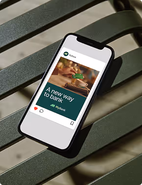 Smartphone on green bench displaying a social media post about MyBank with text 'A new way to bank' and a photo of hands holding a credit card and payment terminal.