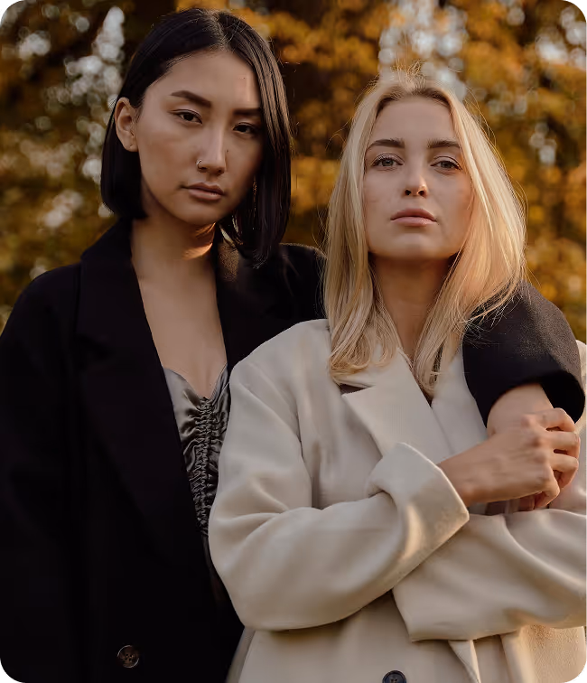 Two women standing close together outdoors with autumn foliage in the background, one with short dark hair wearing a dark coat and the other with long blonde hair wearing a light coat.