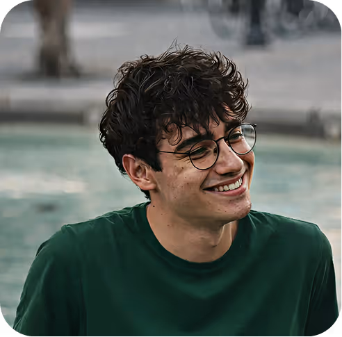 Young man with curly dark hair and glasses smiling outdoors near a fountain.