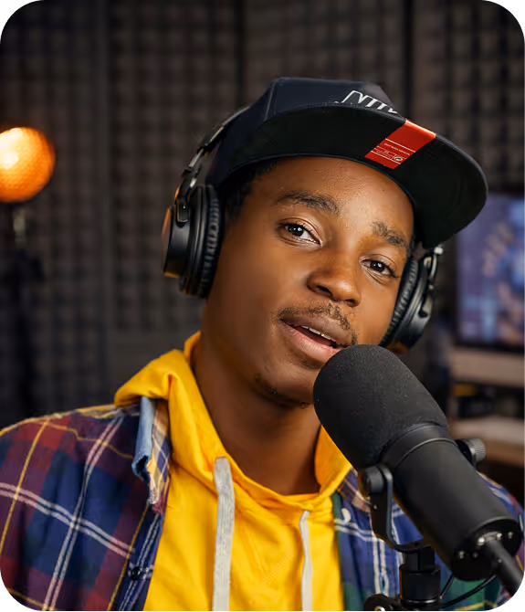 Young man wearing headphones and a black cap speaking into a microphone in a recording studio.