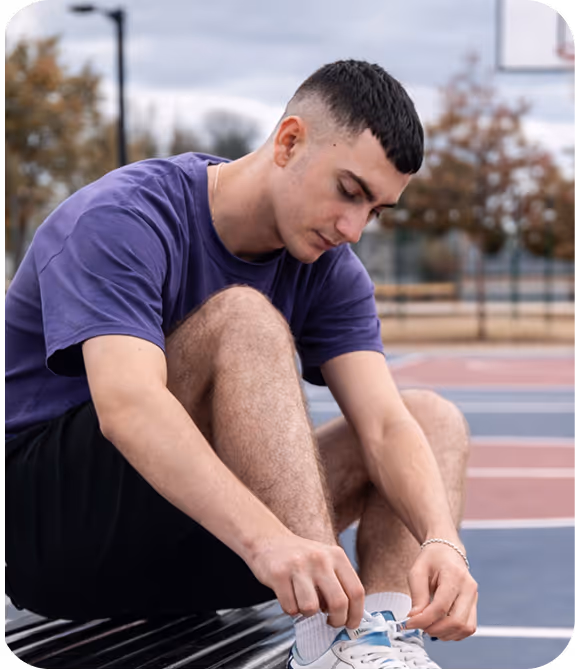 Young man in a purple shirt sitting on a bench tying his shoelaces at an outdoor basketball court.