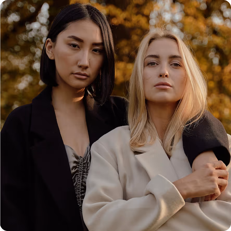 Two women standing closely outdoors with autumn foliage in the background, one with black hair and nose ring wearing a black coat, the other with blonde hair wearing a light-colored coat.