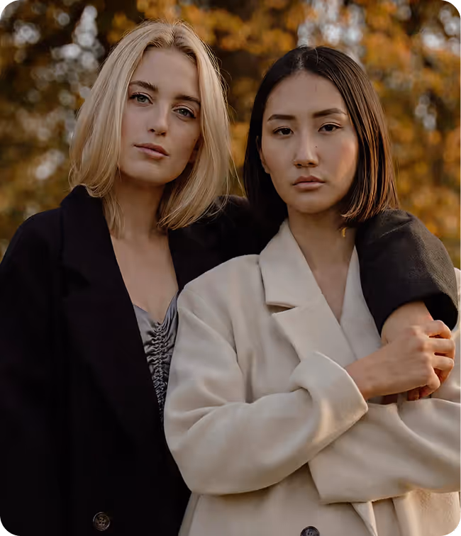 Two women standing outdoors with autumn foliage in the background, one with blonde hair wearing a black coat and the other with straight dark hair wearing a cream-colored coat.