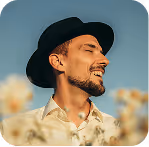 Smiling man with beard and mustache wearing a black hat and white shirt outdoors with blurred flowers.