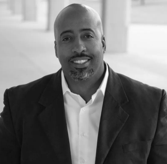 Professional headshot of a confident sales and leadership expert, wearing a dark blazer and white shirt, smiling against an urban background, representing sales coaching and executive leadership themes.