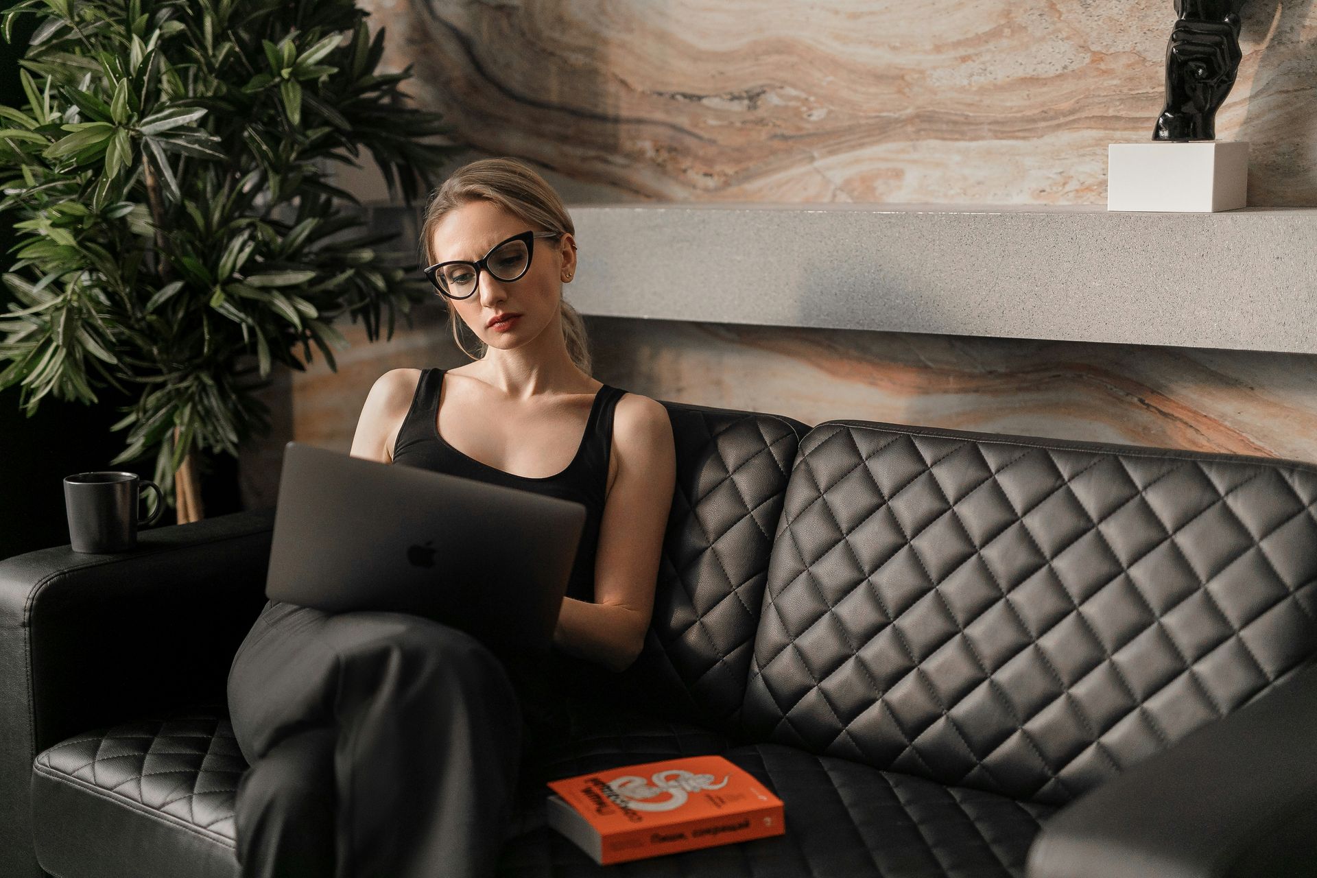 Focused woman working remotely on a laptop while sitting on a black leather couch, representing fractional VP of sales business strategy and flexibility in modern consulting work.