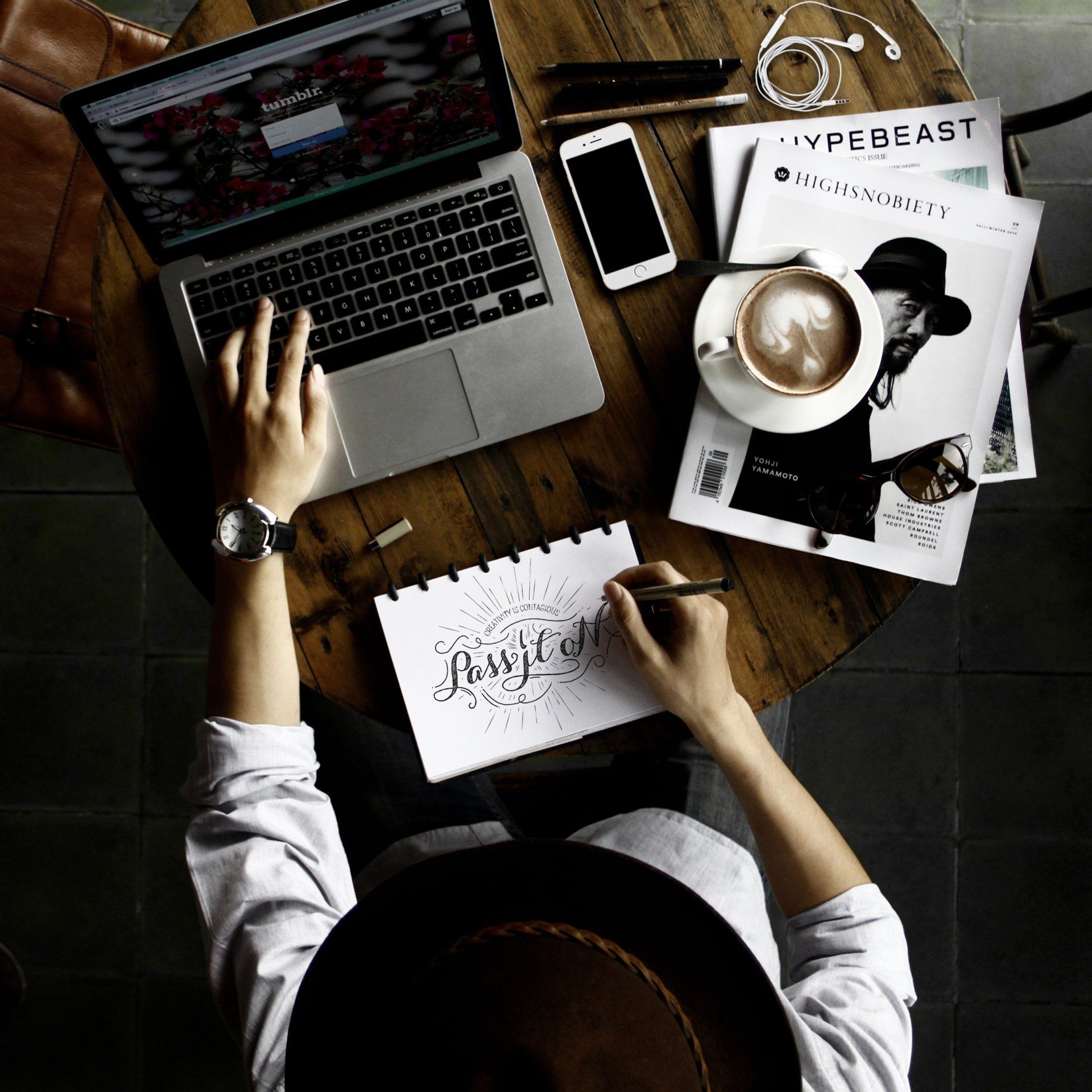 Overhead view of a creative workspace with a person sketching in a notebook while using a laptop. The wooden table is scattered with a smartphone, magazines, a cup of coffee with latte art, and earphones, creating a modern and stylish work environment.