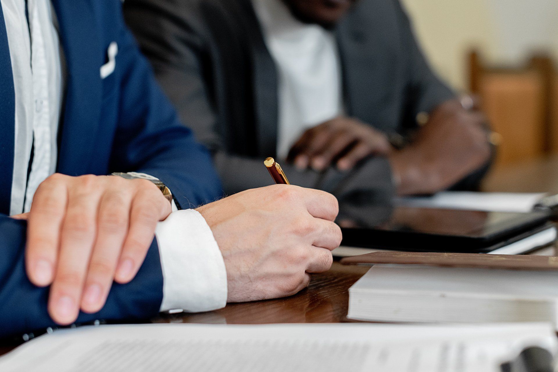 Close-up of two business professionals sitting at a table, one writing notes with a pen while the other rests their arms on the desk, with documents and a tablet in view.