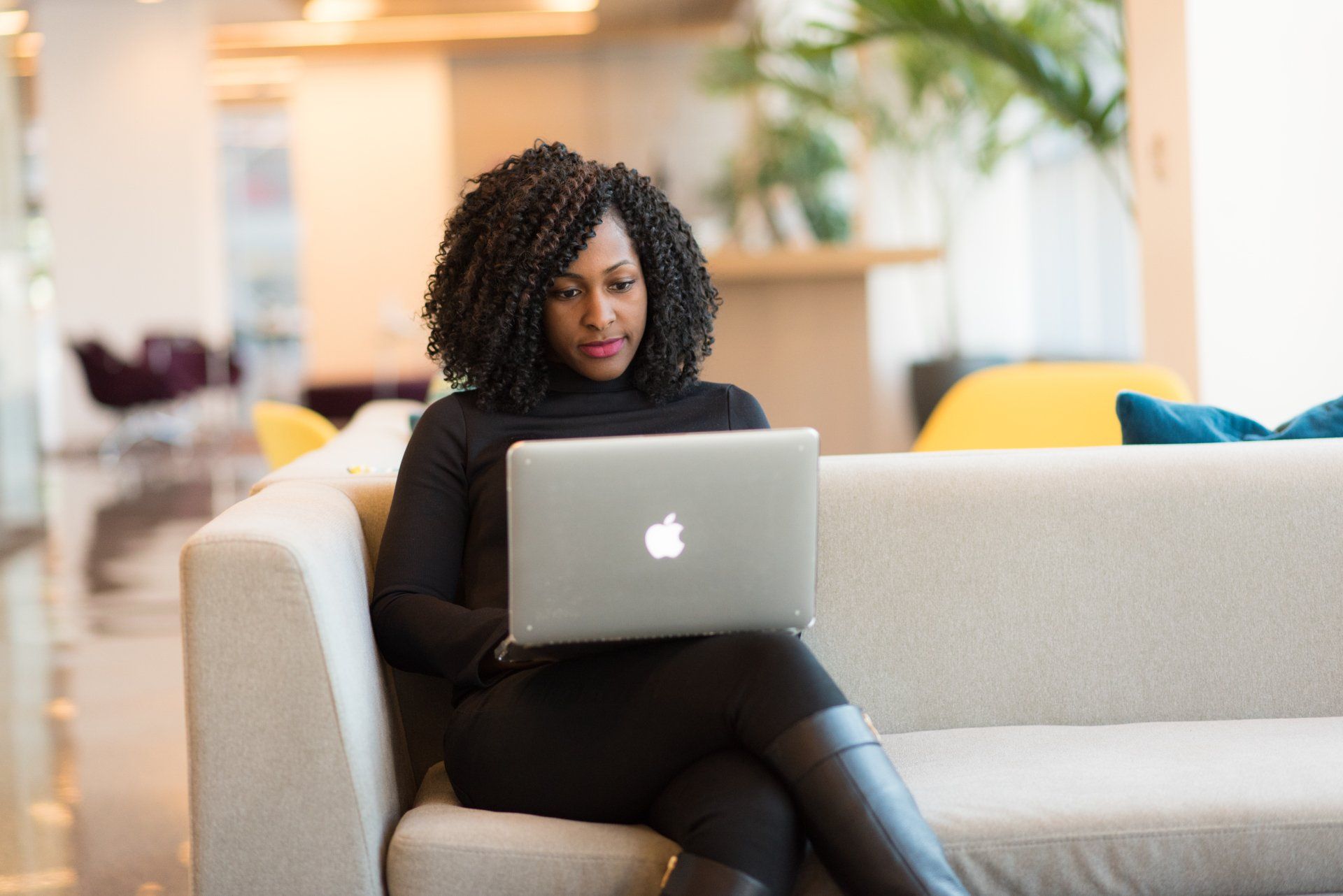 A professional woman sitting on a couch, working on her laptop in a modern office setting.