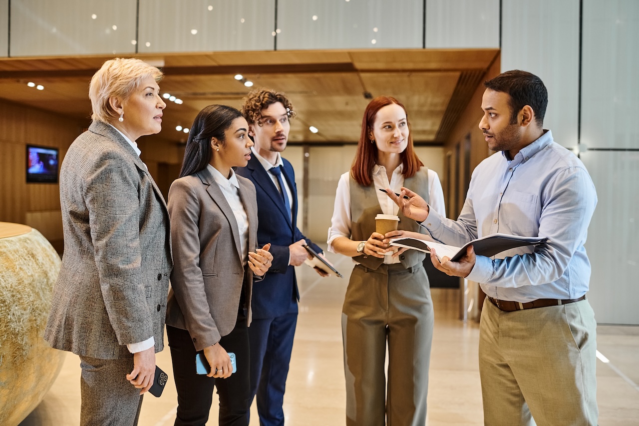 Group of business professionals having a conversation in a modern office lobby, illustrating the collaborative and flexible nature of fractional sales leadership promoted by Revenue Nomad.