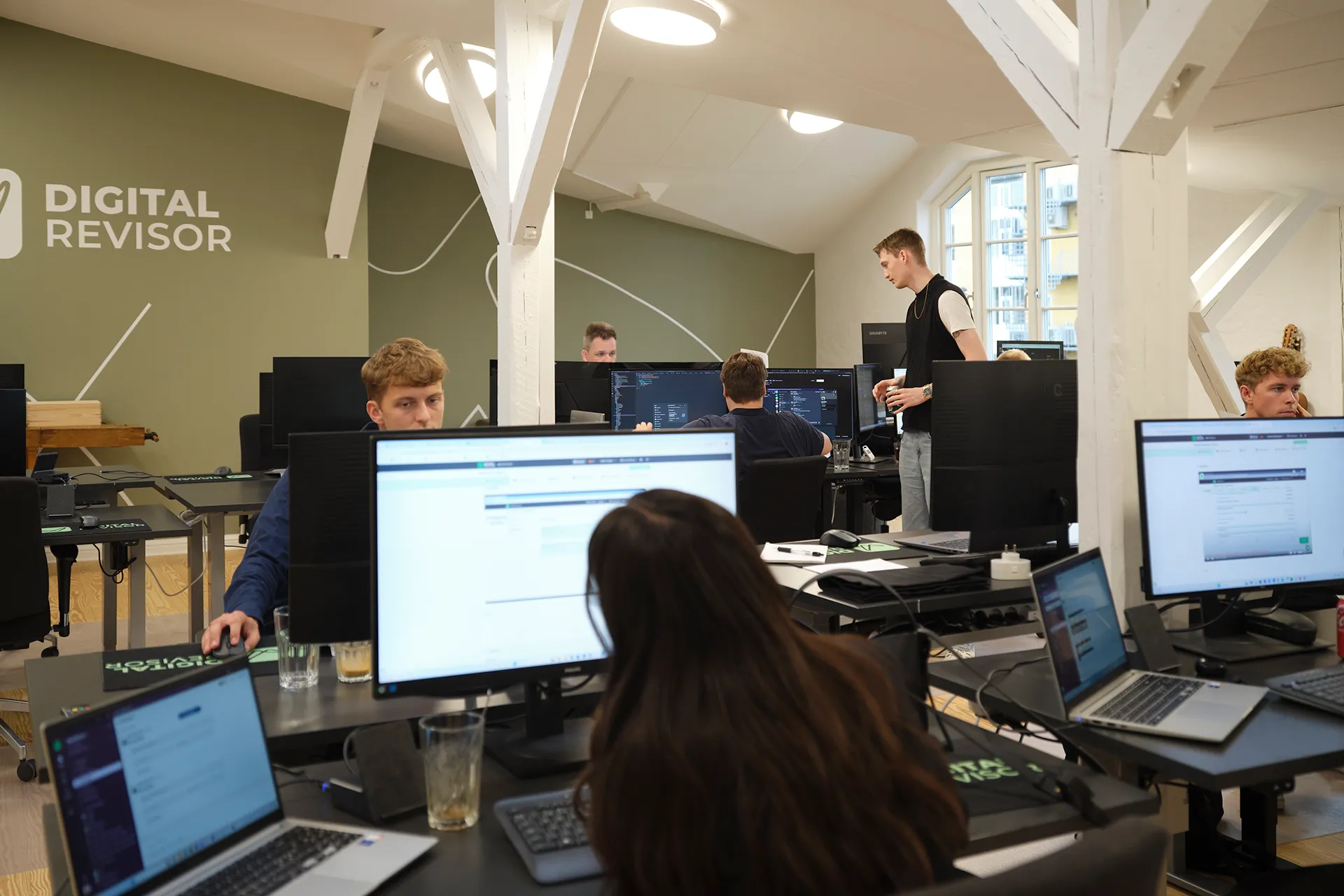 Office workspace with several people working at computers, visible DIGITAL REVISOR logo on green wall.