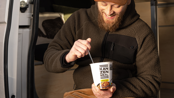 Person sitzt in der geöffneten Tür eines Campingvans, hält einen KANZEN MEAL CUP in der Hand und blickt nach draußen.