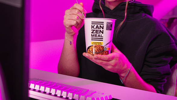 Person sitzt vor einem Computerarbeitsplatz, hält einen KANZEN MEAL CUP in der Hand und blickt auf einen Monitor. Sichtbar sind die Tastatur und die Rückseite des Bildschirms