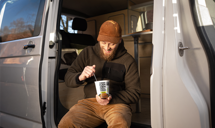 Person sits in the doorway of a camper van holding a KANZEN MEAL CUP while enjoying a meal on the go.