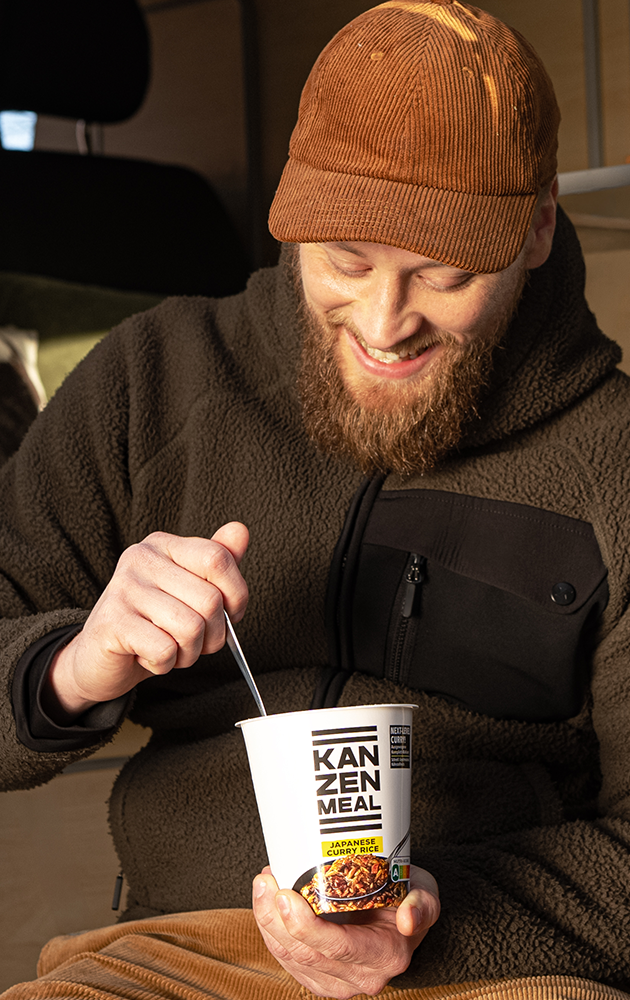 Person sitzt in der geöffneten Tür eines Campingvans, hält einen KANZEN MEAL CUP in der Hand und blickt nach draußen.