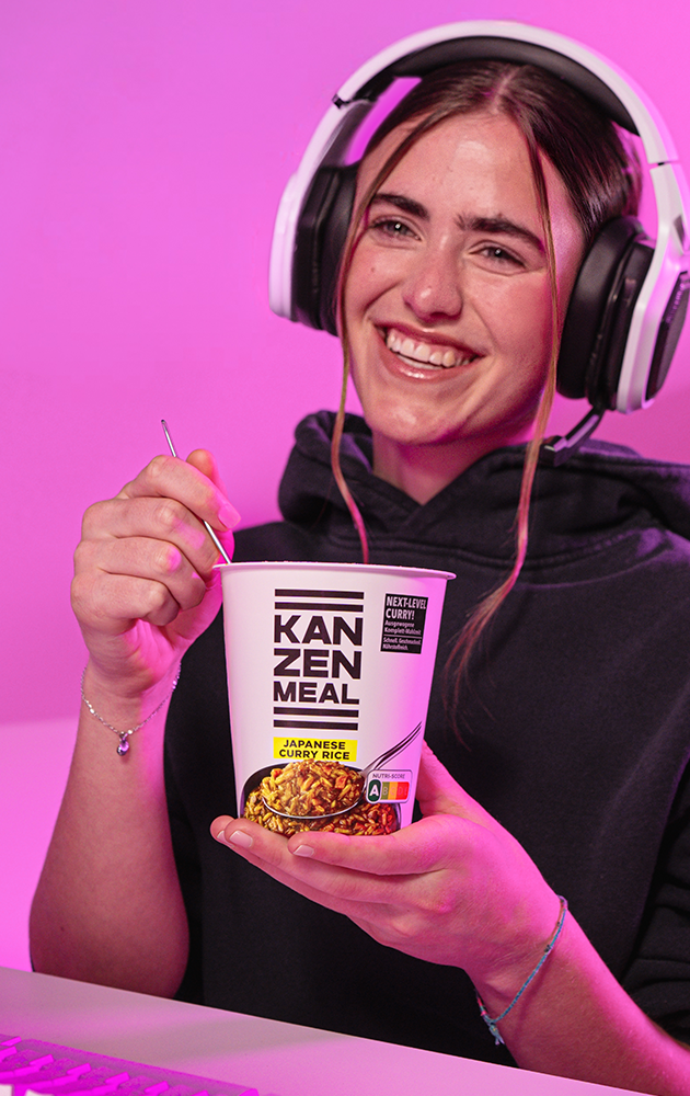 Person sitzt vor einem Computerarbeitsplatz, hält einen KANZEN MEAL CUP in der Hand und blickt auf einen Monitor. Sichtbar sind die Tastatur und die Rückseite des Bildschirms