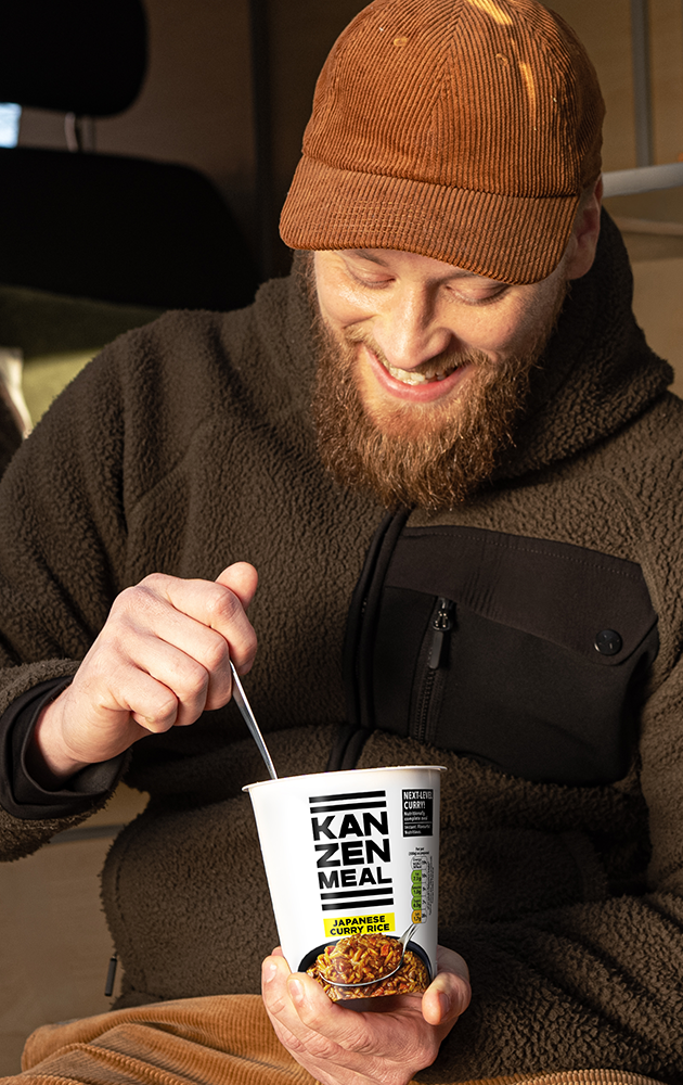 Person sits in the doorway of a camper van holding a KANZEN MEAL CUP while enjoying a meal on the go.