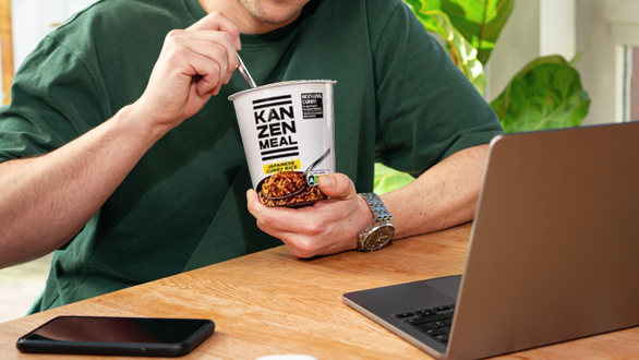 Person sitzt an einem Tisch, hält einen KANZEN MEAL CUP in der Hand und isst. Ein geöffneter Laptop steht vor der Person, daneben liegt ein Smartphone