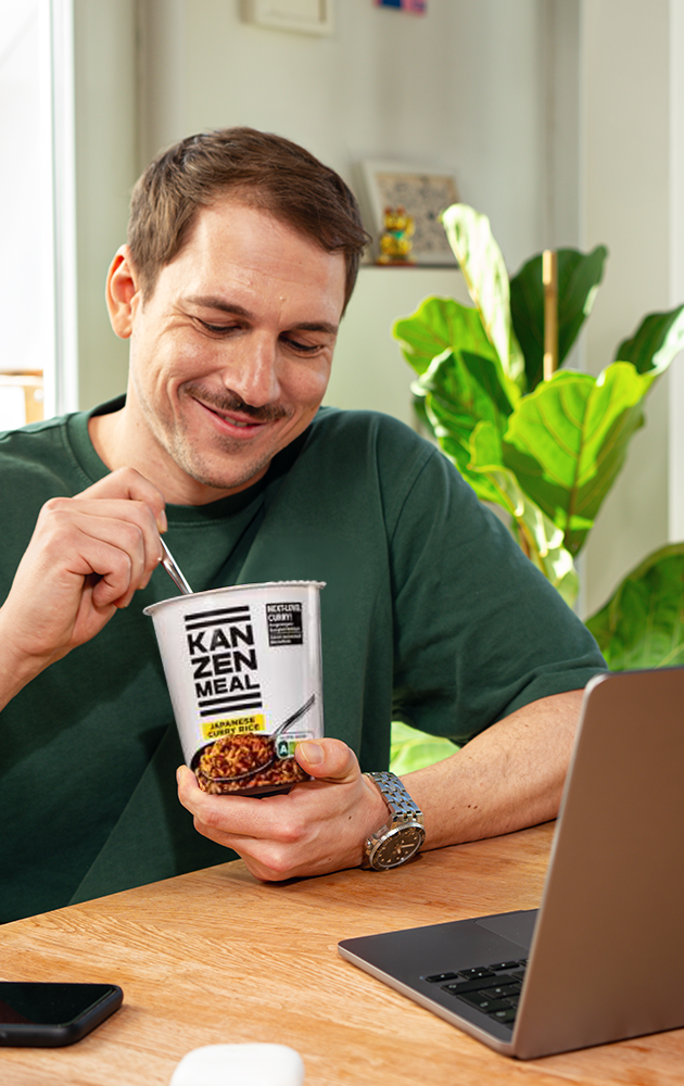 Person sitzt an einem Tisch, hält einen KANZEN MEAL CUP in der Hand und isst. Ein geöffneter Laptop steht vor der Person, daneben liegt ein Smartphone