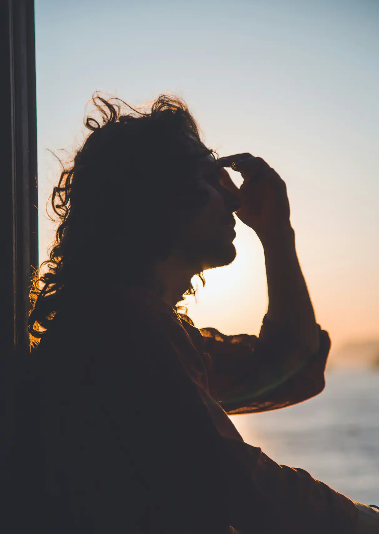 With a low sun in the background, a person sits in the window with their hand softly pinching their forehead