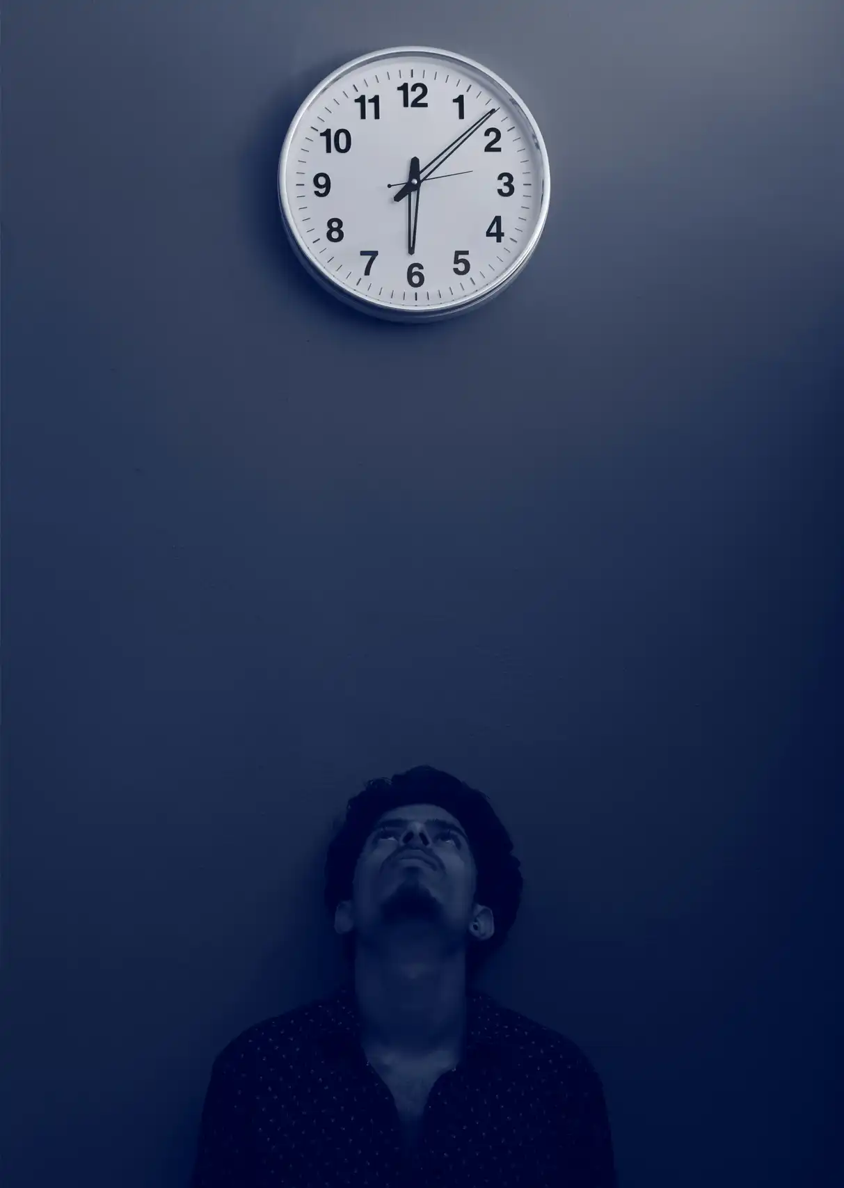 A man in a semi-dark room with his back to the wall looking up at a clock