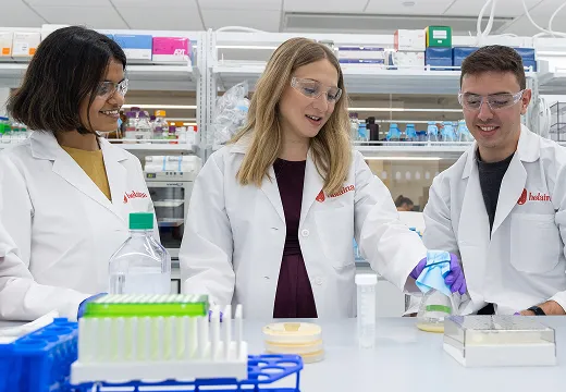 Three scientists in lab coats and safety glasses are working together in Helaina's laboratory.