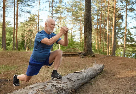 An older man in athletic clothes is doing a lunge exercise in a forest.