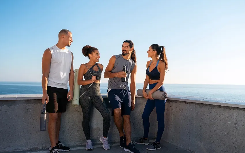 Four friends in workout clothes stand together, smiling and talking after a run by the ocean.