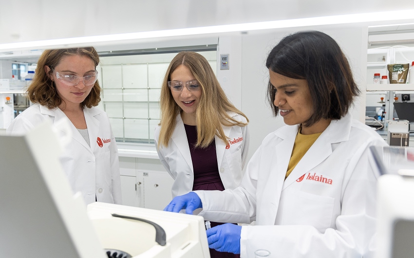 Three women in lab coats look at a machine in Helaina's lab.