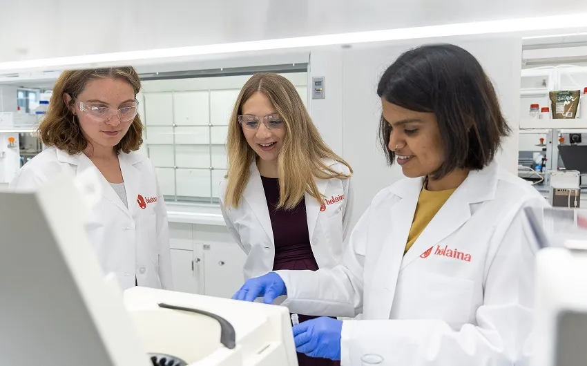 Three women in lab coats look at a machine in Helaina's lab.