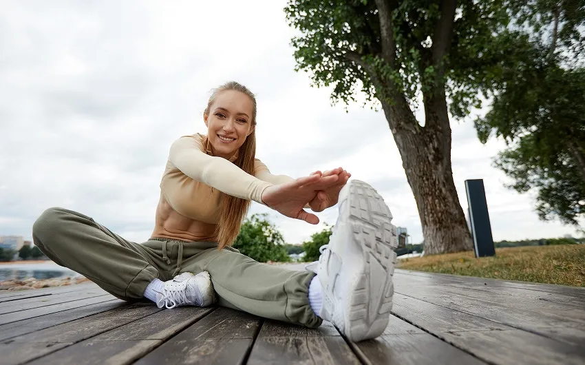 A woman in athletic wear sits on a wooden deck stretching, looking at the camera and smiling.