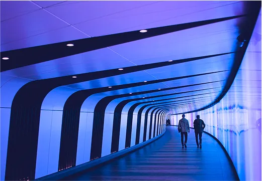 Two people walk away from the viewer down a long, curved, and futuristic-looking hallway with bright blue and purple neon lights.
