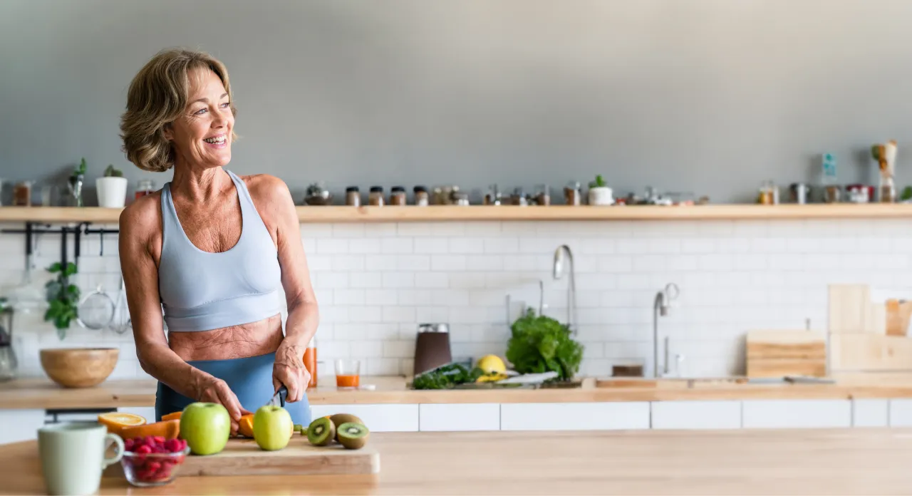 A smiling woman in a sports bra chops fruit in a bright kitchen.